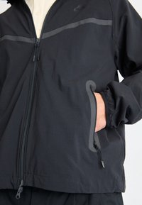 Svart zip-up jacka med huva, med en horisontell grå rand och elastiska ärmar. Innehåller sidofickor och har en mjuk, lätt textur.