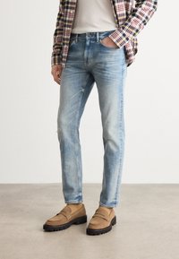 Light blue denim jeans with a straight-leg fit, featuring subtle distressing. Paired with brown loafers and a checkered shirt layered over a t-shirt.