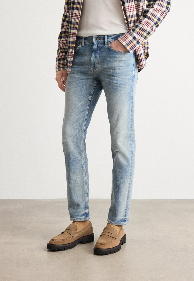 Light blue denim jeans with a straight-leg fit, featuring subtle distressing. Paired with brown loafers and a checkered shirt layered over a t-shirt.