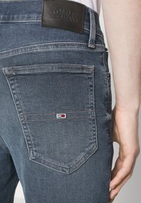 Tmavě modré džínové kalhoty s koženou záplatou na pásu označenou "TOMMY JEANS." Mají dvě zadní kapsy s malým barevným logem.