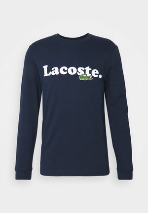 Marineblå langærmet t-shirt lavet af bomuld, med hvid "Lacoste." tekst og en broderet grøn krokodillelogo på venstre side.