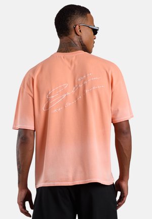 Mann trägt Sonnenbrille und pfirsichfarbenes T-Shirt mit weißer kursiver Schrift auf dem Rücken, kombiniert mit schwarzen Shorts, schaut von der Kamera weg.