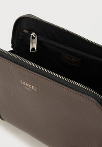 Sötétbarna bőr zipzáras pénztárca texturált felülettel. Fekete szövet belsővel és kis cipzáras zsebbel rendelkezik. A Lancel logó az elején gravírozott.