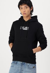 Sudadera negra con bolsillo tipo canguro, que presenta un logo blanco en el pecho. Hecha de algodón, tiene capucha con cordón y puños acanalados.