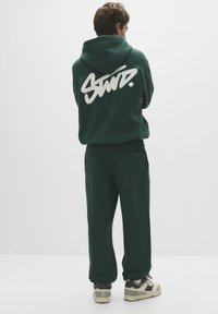 Hanorac verde cu glugă, având un logo mare alb "STWD." pe spate. Asortat cu pantaloni verzi și pantofi negri/aiuri. Textură netedă.