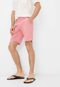 Pantaloni corti rosa in cotone con una vestibilità dritta, indossati con una camicia bianca in lino e infradito nere. Il tessuto ha una sottile trama.