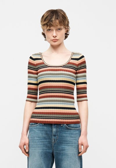 Jeune personne aux cheveux courts et bouclés, portant un haut côtelé ajusté à rayures multicolores horizontales et un jean bleu, sur fond uni.