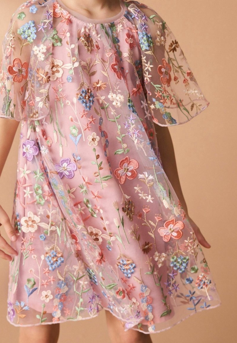 Enfant portant une robe rose en voile avec des fleurs colorées brodées et des manches papillon sur un fond beige uni.