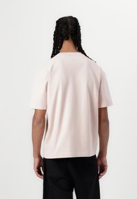 Svetlo roza kratka majica s kratkimi rokavi, sproščenega kroja, okroglim ovratnikom in gladko teksturo. Zadnji del prikazuje minimalen dizajn in čiste linije.