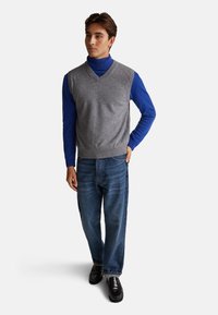Szary sweter bez rękawów na niebieskim golfie, zestawiony z niebieskimi dżinsami i czarnymi butami. Sweter ma dekolt w kształcie litery V i prążkowane wykończenia.