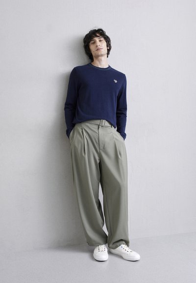 Chemise à manches longues bleu marine avec col côtelé, associée à un pantalon plissé vert clair et des baskets blanches, se tenant devant un fond neutre.