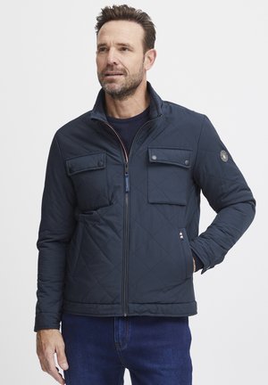 Marineblaue Steppjacke mit hohem Kragen, zwei knöpfbaren Brusttaschen, seitlichen Taschen und Logopatch am linken Ärmel. Glatte Textur.