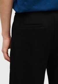 Κοντινό πλάνο ενός ατόμου που φοράει μαύρο παντελόνι με τσέπη στην πίσω πλευρά και μπλε μπλούζα, εμφανίζοντας τον κάτω κορμό και το αριστερό χέρι.