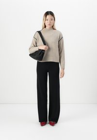 Donna che indossa un maglione beige, pantaloni neri a gamba larga, scarpe rosse con punta, mentre tiene una borsa nera a spalla su uno sfondo bianco semplice.