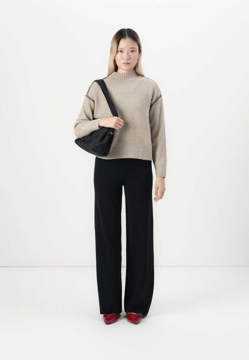 Frau in beigem Pullover, schwarzer weit geschnittener Hose, roten spitz zulaufenden Schuhen, die eine schwarze Umhängetasche vor einem schlichten weißen Hintergrund hält.