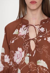 Bruine bloemenblouse met een geknoopt decolleté, van lichtgewicht stof en een losse pasvorm. Bloemenpatronen in roze en groene accenten zorgen voor detail.