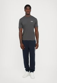 Grå kortermet t-skjorte med logo på venstre bryst, kombinert med marineblå joggebukser med hvit tekst, og hvite sneakers.