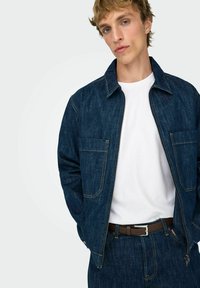 Temno modra jeans jakna z zadrgo spredaj, dvema velikima žepoma in kontrastnim šivom. Nošena preko bele majice in rjavega pasu.