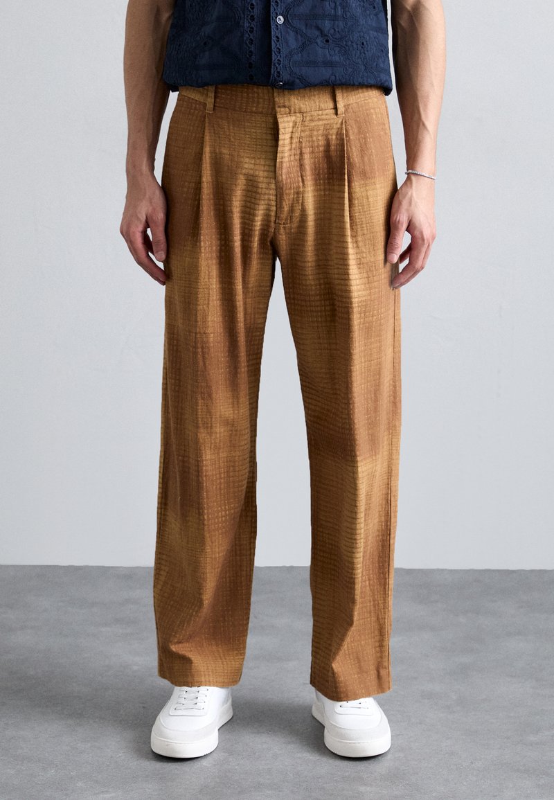 Brown plaid trousers with a textured fabric, featuring a loose fit and front pleats, paired with white sneakers.