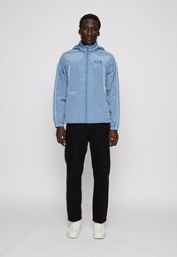 Světle modrá, lehká, kapucí vybavená bunda se zipem, elastickými manžetami a bílými bočními pruhy, kombinovaná s černými kalhotami a bílými teniskami.