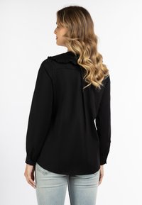 Schwarze Bluse mit langen Ärmeln, die einen Rüschensaum und einen abgerundeten Saum aufweist, aus einem glatten Stoff gefertigt. Das Model trägt hellblaue Jeans.