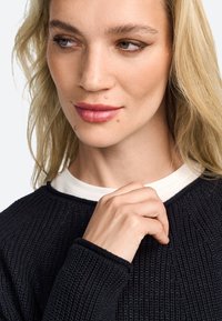 Maglione lavorato a maglia blu navy con texture a coste, dotato di scollo rotondo. Un colletto interno bianco è visibile e la mano del modello poggia sul tessuto.