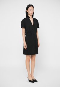 Μαύρο φόρεμα με ρίγες, βαθύ V-ντεκολτέ, κοντά μανίκια και εφαρμοστή μέση με κλείσιμο από κουμπί. Συνδυάζεται με μαύρες μυτερές γόβες.