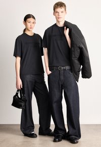 Tricouri negre din bumbac cu logo embosat, asortate cu pantaloni largi din denim închis la culoare. Un model ține o geantă mică neagră; jachetă neagră structurată.