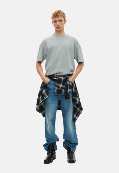 Camiseta de gran tamaño color gris claro, jeans de mezclilla azul, camisa de cuadros negra atada a la cintura, botas negras. Textura suave, corte relajado.
