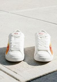 Sneakers bianche con disegni di fiamme rosse e gialle ai lati, posizionate fianco a fianco su una superficie di cemento.
