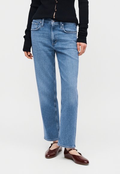 Άτομο που φοράει ανοιχτό μπλε τζιν παντελόνι με ίσια γραμμή, μαύρο ζακέτα με κουμπιά και σκοτεινά κόκκινα παπούτσια Mary Jane, στέκεται σε λευκό απλό φόντο.