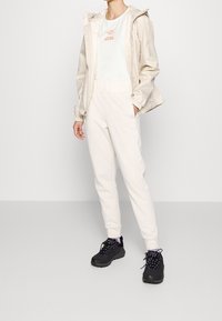 Giacca impermeabile beige con cappuccio, abbinata a una maglietta chiara e pantaloni jogger color crema. Sneakers nere con suole testurizzate completano l'outfit.