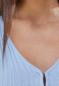 Top in maglia a costine di un blu chiaro, con profondo scollo a V e una piccola chiusura zip frontale. Texture liscia con linee verticali ben visibili.