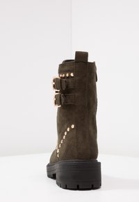 Bottine en daim vert olive avec double boucle et clous dorés sur le côté, semelle en caoutchouc noir et talon plat.