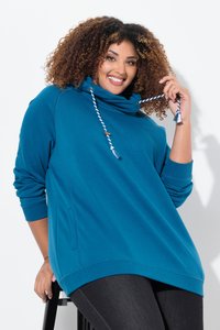 Femme souriante aux cheveux bouclés portant un sweat à capuche bleu avec des cordons, assise sur un tabouret noir sur fond blanc.
