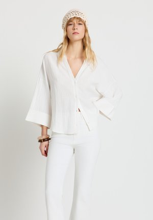 Femme portant un chemisier blanc à boutons, un pantalon blanc, un bonnet en crochet beige et des bracelets, posant devant un fond clair uni.