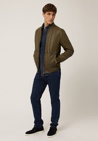 Olivgrüne Bomberjacke aus Leder mit gerippten Bündchen und Saum, kombiniert mit einem Denimhemd, dunklen Jeans und schwarzen Sneakern.