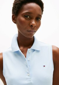 Polo sans manches bleu clair avec un col pointu, une patte de boutonnage à deux boutons et un petit logo brodé sur la poitrine. Tissu texturé.