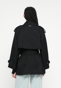 Czarny trencz o krótkim kroju, z pagonami i paskiem w talii. Posiada subtelne logo z tyłu oraz gładką fakturę tkaniny.