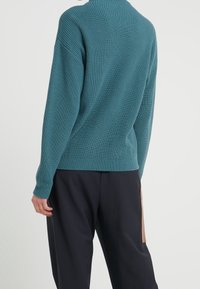 Maglione a coste color teal con collo alto e maniche lunghe, caratterizzato da un motivo a maglia testurizzata. Indossato con pantaloni neri larghi.