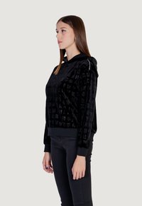 Sudadera de terciopelo negro con patrones de logotipos en relieve, capucha con cordón y puños de canalé. Combinada con pantalones negros.