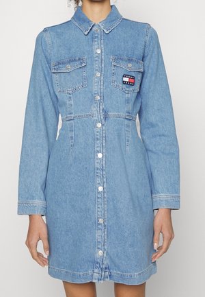 Femme portant une robe chemise en denim bleu clair avec manches longues, deux poches poitrine, et un patch logo Tommy Jeans sur la poche gauche.