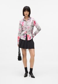 Srajca s gumbi v roza in sivi barvi z abstraktnimi vzorci, v kombinaciji s črno nabrano krilo. Dodatki vključujejo majhno črno torbico in pete.