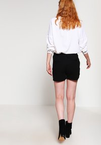 Schwarze Shorts mit Bund und Gesäßtaschen, kombiniert mit einem weißen langärmligen Oberteil. Modell trägt schwarze Stiefel mit Absatz.