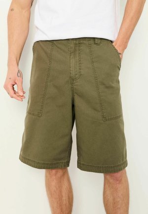 Mann trägt olivgrüne knielange Shorts mit aufgesetzten Fronttaschen und ein weißes T-Shirt, steht mit einer Hand in der Tasche.