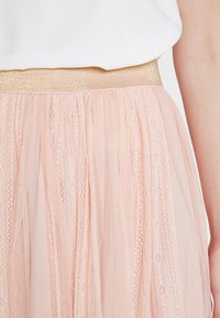 Valkoinen paita on sujautettu vaaleanpunaiseen pitsihameeseen, jossa on kimalteleva kultainen joustava vyötärö, ja kuva näyttää henkilön käsivarren sivusta.