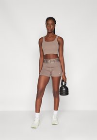 Sujetador deportivo marrón y pantalones cortos con cinturilla "NIKE PRO". Lleva calcetines blancos y zapatillas ligeras, sosteniendo una kettlebell negra. Textura suave.