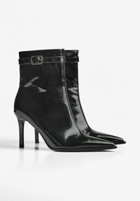 Musta patentoitu nahka nilkkurit, joissa on terävä kärki, stiletto-korko ja koristeellinen hihna, jossa on hopeinen solki.