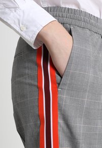 Šedé kostkované kalhoty s elastickým pasem, obsahující boční kapsu a pruhované akcenty v červené, bílé a burgundské barvě podél nohavic.