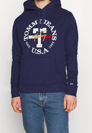 Osoba ubrana w granatową bluzę z kapturem Tommy Jeans z białym napisem logo i dżinsami, stoi na tle jasnego, jednolitego tła.